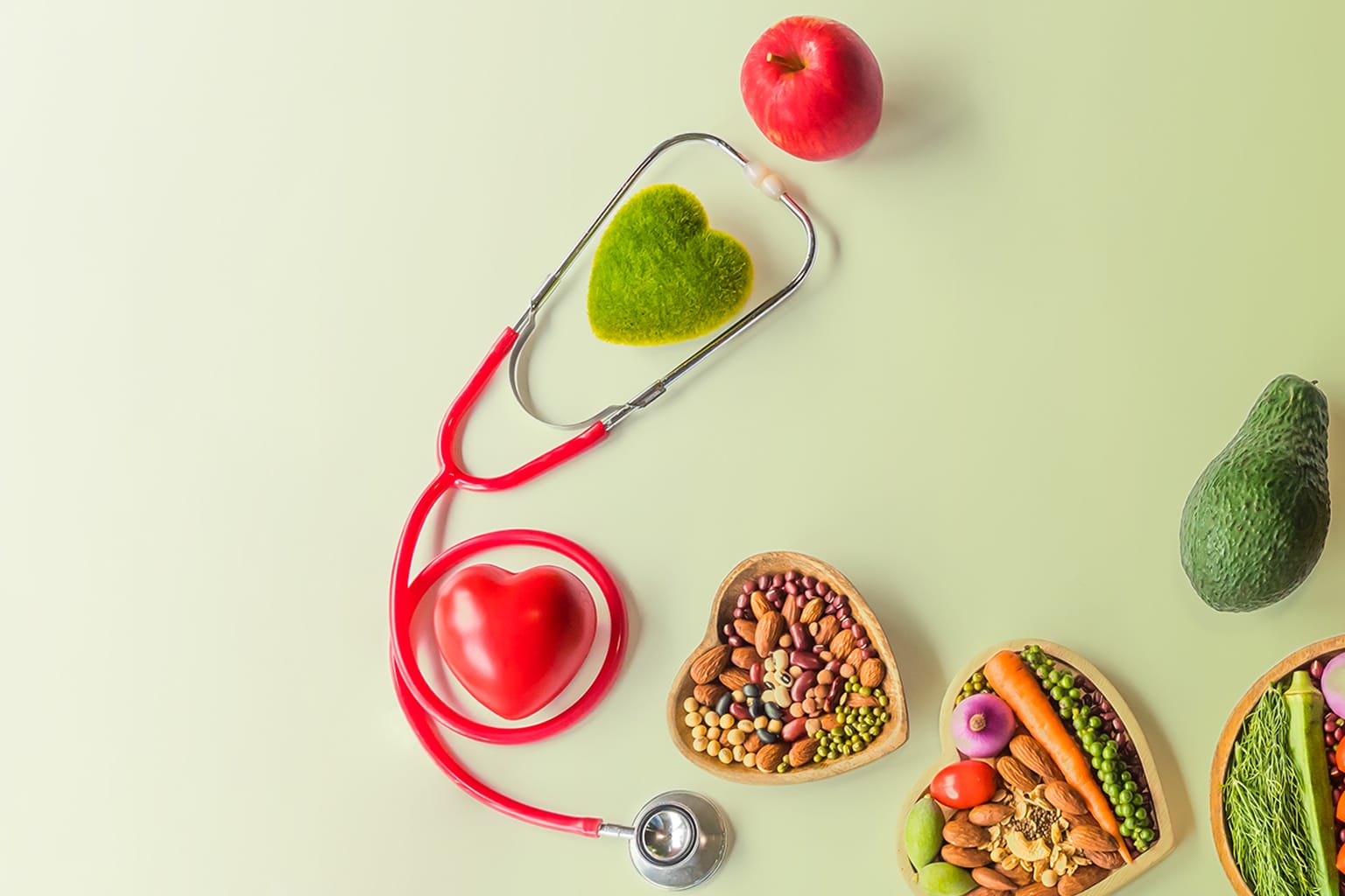 Uno stetoscopio rosso su sfondo verde chiaro, circondato da frutta, verdura e legumi in bacinelle a forma di cuore.